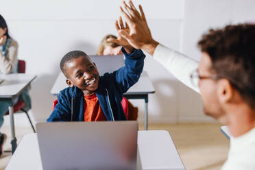 Happy black kid doing a high five with his teacher in a computer science classroom, celebrating a successful teaching and learning moment. Young student enjoying individual attention and assistance from his teacher as he learns digital literacy. - JLPSF29717