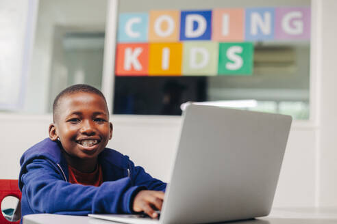 Excited young boy smiling at the camera as he sits with a laptop in a coding classroom. Young black student practicing his programming skills in a computer-based learning school. - JLPSF29731