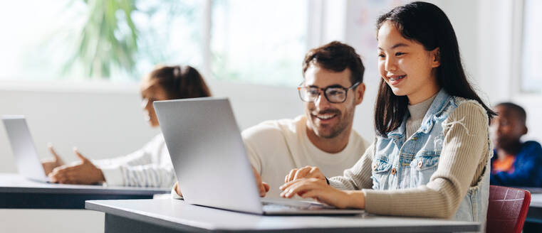 Happy young girl getting assistance from her teacher as she engages in a computer science lesson. Female student learning and understanding the basics of digital literacy in a classroom. - JLPSF29742