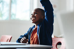 Ein kleiner schwarzer Junge hebt die Hand, um eine Frage in einem Grundschulklassenzimmer zu beantworten. Mit einem zuversichtlichen Lächeln im Gesicht nimmt der Junge am Unterricht teil, der von seinem Lehrer geleitet wird. - JLPSF29748
