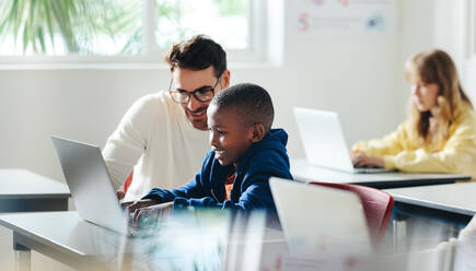 Ein männlicher Lehrer hilft einem Jungen beim computergestützten Lernen in einem Klassenzimmer. Ein Tutor unterrichtet ein Kind in einer Grundschule, wobei der Schwerpunkt auf Programmieren und digitalen Grundkenntnissen liegt. - JLPSF29751