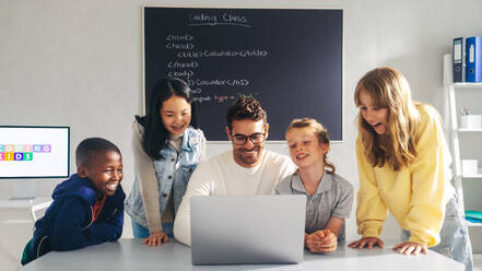 Ein Lehrer der Coding School zeigt einer Gruppe von Schülern, wie man auf einem Laptop programmiert. Glückliche Kinder lernen digitale Kompetenz und erwerben eine solide Grundlage in Informatik, während sie sich mit ihrem Lehrer in einer Klasse beschäftigen. - JLPSF29766