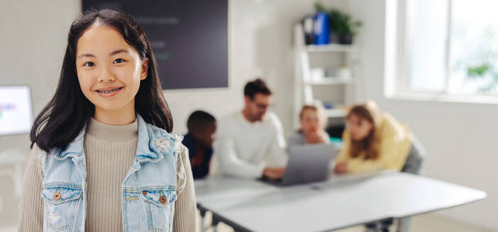 A confident student looks at the camera while the teacher engages other students in the background. Girl attending a digital literacy class at school. - JLPSF29768