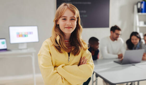 Female student standing in a tech education classroom, looking the camera, while her teacher and classmates code in the background. Confident young girl looking ready to take on a programming lesson. - JLPSF29771