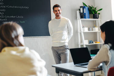 Junger männlicher Lehrer, der einen Programmierkurs für Kinder leitet. Informatiklehrer, der mit einem Lächeln vor der Klasse steht und die Kinder auf dem Weg in eine bessere Zukunft mit technischen Kenntnissen anleitet. - JLPSF29776