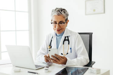 Female general practitioner using a smartphone at her desk. Mature doctor using smart medical apps to manage her patient schedules. Tech-savvy doctor working in her office, wearing a white lab coat. - JLPSF29780