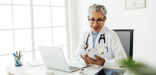 Doctor in a lab coat smiling as she uses a smart app on her mobile phone. Mature female physician working with technology to access medical information and connect with her patients in her office. - JLPSF29781
