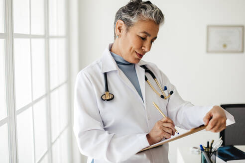 Mature female doctor writing patient information on a clipboard in her office, wearing a white lab coat. Experienced health professional documenting treatment plans for a patient after an appointment. - JLPSF29782