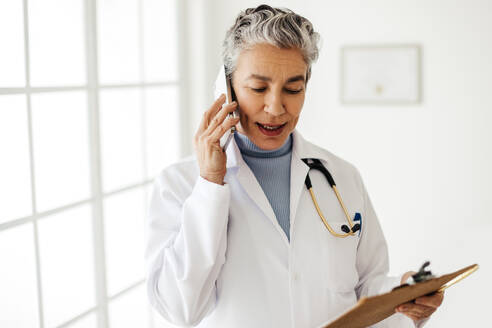 Doctor speaking to a patient on a phone call, discussing their test results. Mature female health professional explaining her diagnosis to a patient over the phone, as she stands with a clipboard in her hand. - JLPSF29783