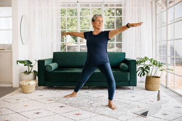 Mature woman showing her strength and flexibility as she practices the powerful warrior pose at home. Active senior woman doing yoga in an online fitness class, following a healthy workout routine. - JLPSF29788