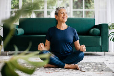 Glückliche ältere Frau, die Yoga im Lotussitz praktiziert und für inneren Frieden und Achtsamkeit meditiert. Ältere Frau, die eine Atemübung in der Ruhe ihres Hauses macht, in Fitnesskleidung auf dem Boden sitzend. - JLPSF29789