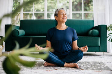 Ältere Frau, die in ihrem Wohnzimmer Yoga praktiziert, im Lotussitz sitzt und meditiert. Diese glückliche ältere Frau genießt die Ruhe und Gelassenheit dieser Atemübung, wie das Lächeln auf ihrem Gesicht zeigt. - JLPSF29790