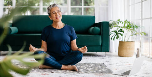 Ältere Frau übt Yoga in einem Online-Fitnesskurs, sie sitzt auf dem Boden und meditiert im Lotussitz vor einem Laptop. Glückliche ältere Frau, die zu Hause ein Trainingsprogramm für Entspannung und Achtsamkeit macht. - JLPSF29791