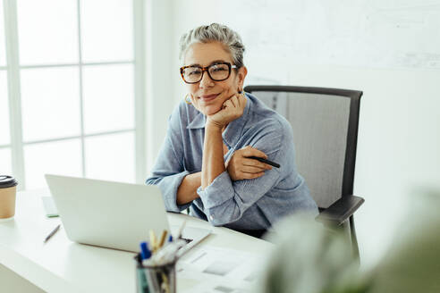 Senior-Grafikdesignerin, die mit einem Stift in der Hand in die Kamera blickt, während sie in ihrem Büro an einem anderen Projekt arbeitet. Diese Frau widmet sich voll und ganz ihrem Handwerk und erstellt Designs mit einem Laptop. - JLPSF29809