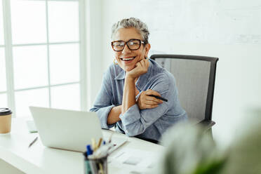 Happy senior woman looking at the camera with a smile, enjoying working on a graphic design project in her office. Successful female professional working with a laptop and a graphics tablet to perfect her craft. - JLPSF29810