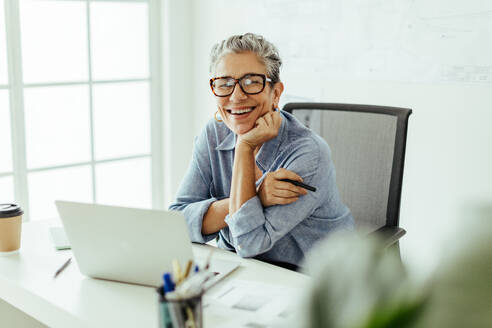 Glückliche ältere Frau, die lächelnd in die Kamera blickt und sich an einem Grafikdesign-Projekt in ihrem Büro erfreut. Erfolgreiche weibliche Fachkraft, die mit einem Laptop und einem Grafiktablett arbeitet, um ihr Handwerk zu perfektionieren. - JLPSF29810