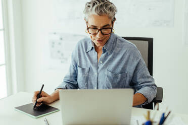 Senior Designerin, die ein Grafiktablett und einen Laptop benutzt, um ihre Entwürfe zum Leben zu erwecken und visuelle Meisterwerke zu schaffen. Erfahrene Fachfrau, die mit technischen Hilfsmitteln arbeitet, um ihr Handwerk in ihrem Büro zu perfektionieren. - JLPSF29812