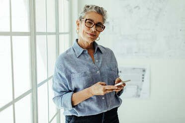 Portrait of a professional woman standing in her office and using a mobile phone. Senior design expert looking at the camera with confidence as she types a text message to a client. - JLPSF29820