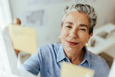 Mature designer using sticky notes to lay out her ideas in an office. Experienced business woman planning a project, writing out her plans on a glass wall. Senior female architect at work. - JLPSF29826