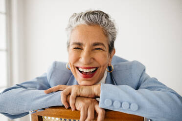 Business woman with grey hair expressing pride in her career success and accomplishments. Female business expert smiling with joy as she sits in her office dressed in a professional suit. - JLPSF29831