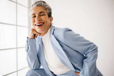 Successful business woman smiling at the camera, confident and empowered in her field as an experienced professional. Business woman with silver hair standing in her office, dressed in a classy suit. - JLPSF29835