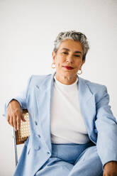 Mature business woman embodying confidence and empowerment as she sits on an office chair in a professional suit. Senior female executive looking at the camera with a bold expression, dressed in elegant business attire and silver hair. - JLPSF29836
