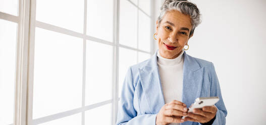 Ältere Geschäftsfrau mit grauen Haaren, die bei der Arbeit ihr Smartphone benutzt. Ältere Geschäftsfrau, die in einem Büro steht und die mobile Technologie benutzt, um mit ihren Geschäftspartnern in Kontakt zu treten. - JLPSF29850