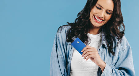 Gen Z woman smiles happily while holding a cred it card in her hand in a studio setting, she embraces the use of mobile and online payments. She enjoys shopping online and paying with a single tap. - JLPPF01627