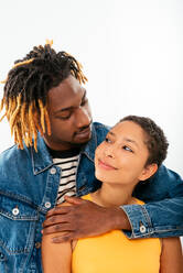 Side view of smiling African American boyfriend with dreadlocks in denim jacket hugging happy girlfriend with short hair looking at each other in studio - ADSF43740