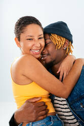 Seitenansicht eines lächelnden afroamerikanischen Freundes mit Dreadlocks in Jeansjacke, der eine glückliche Freundin mit kurzen Haaren in Jeans im Studio umarmt - ADSF43748