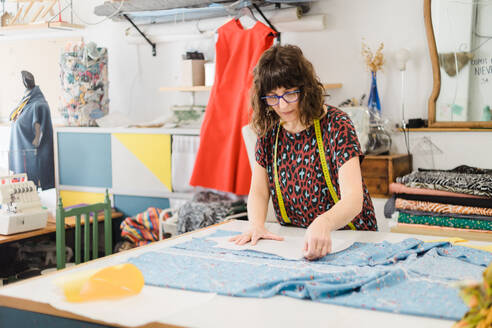 Konzentrierte Designerin in legerer Kleidung und Schal, die ein Muster auf einem Stoff vorbereitet, während sie am Tisch im Atelier arbeitet - ADSF43765