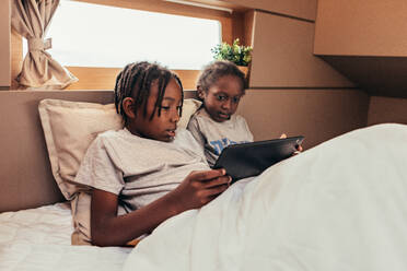 Ruhige schwarze Kinder, die unter einer Decke auf dem Bett liegen und gemeinsam einen Film über ein Tablet ansehen, während sie sich am Wochenende ausruhen - ADSF43772