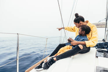Full body of happy African American mother in casual clothes sitting on yacht and pointing away while smiling and showing view to little son during weekend - ADSF43775