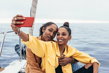 Fröhliche afroamerikanische Freunde umarmen sich und halten Erinnerungen mit ihrem Handy fest, während sie auf einer Yacht entspannen - ADSF43777