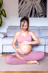 Ganzer Körper einer fröhlichen schwangeren Frau in rosa Aktivkleidung, die lächelt und ihren Bauch berührt, während sie auf einer Yogamatte in Lotus-Pose vor einem Sofa sitzt - ADSF43780