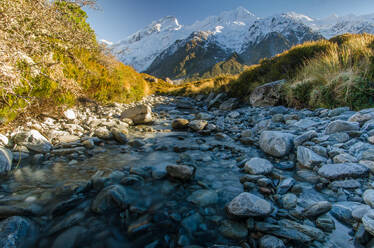 Malerischer Blick auf schneebedeckte Felsen in der Nähe des durch Felsen fließenden Hooker River an einem sonnigen Tag im Aoraki National Park - ADSF43842