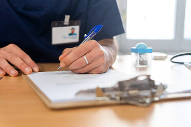 Anonymer Arzt in medizinischer Uniform sitzt am Tisch und schreibt Rezepte auf dem Klemmbrett, während er in einem modernen Krankenhaus arbeitet - ADSF43846