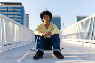 Ganzer Körper eines ernsthaften jungen asiatischen männlichen Skaters in Freizeitkleidung, der in die Kamera schaut, während er auf einem Skateboard auf einer Brücke sitzt - ADSF43879