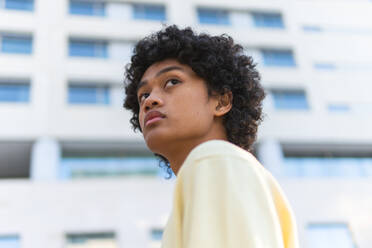 Nachdenklicher junger Mann mit Afrofrisur und gelbem Sweatshirt, der vor einem hohen Gebäude steht und wegschaut - ADSF43883