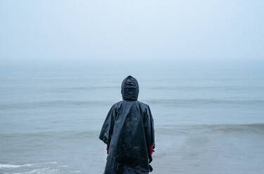 Rückenansicht einer nicht erkennbaren weiblichen Reisenden im Regenmantel, die am Meeresufer steht und das stürmische Wetter unter dem Himmel bewundert - ADSF43885