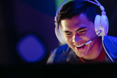 Ein Spieler lächelt auf seinen Computerbildschirm und genießt seine Erfahrung bei einem Online-Spiel. Ein glücklicher junger Mann sitzt an einem PC mit einem Headset und streamt ein Videospiel auf einer Online-Plattform. - JLPSF29865