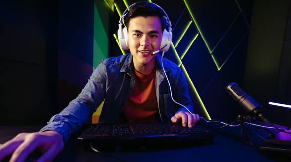 Ein glücklicher junger Mann sitzt mit einem Headset an einem Gaming-Computer und spielt ein rasantes Ego-Shooter-Videospiel. Mit einem Mikrofon in der Nähe streamt er live und bringt das Spielerlebnis zu den Zuschauern eines Online-Streaming-Dienstes. - JLPSF29868