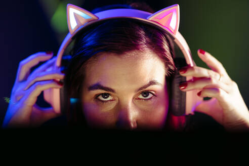 Ein Mädchen setzt ihr Headset auf und bereitet sich auf ein nächtliches Online-Videospiel vor. Eine Gamerin, die in die Welt des Esports einsteigt, sitzt vor einem leuchtenden Computerbildschirm und sieht dabei sehr engagiert aus. - JLPSF29908