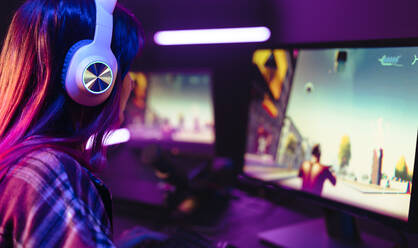 Girl sitting in front of an action-packed computer screen, navigating a first person shooter video game online. Young female gamer engaging with the virtual gaming world with a pc and a headset. - JLPSF29914