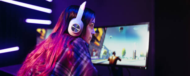 Gamer concentrating on her video game as she sits in front of a multi-monitor setup with a headset. Young woman getting immersed in the realm of digital gaming as she streams an online video game. - JLPSF29916