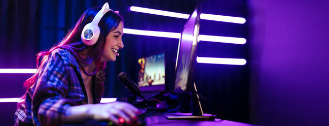 Happy young woman live streaming a video game, sitting in front of a multi monitor setup with a microphone for commentary and a headset for communication. Female engaging with fellow gamers on an online platform. - JLPSF29917
