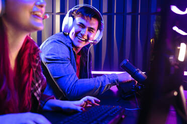 Ein männlicher Spieler lächelt stolz, nachdem er seinen Konkurrenten in einem Online-Gaming-Wettbewerb besiegt hat. Zwei junge Spieler spielen Videospiele auf ihren Computern, wobei ein Mikrofon das Spielgeschehen über einen Live-Streaming-Dienst aufzeichnet. - JLPSF29923