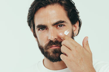 Man looking at the camera as he takes a moment to pamper himself with some quality skincare. Confident man applying some face cream to keep his skin hydrated and healthy, gently massaging it into his face. - JLPSF29957