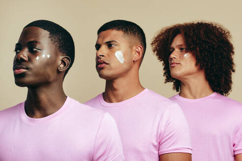 Drei junge Männer mit unterschiedlichen Hauttypen stehen zusammen und schauen selbstbewusst in die Ferne. Eine Gruppe von Männern verwendet eine Schönheitscreme für das Gesicht, um das jugendliche, gesunde Aussehen ihrer einzigartigen Hauttöne zu erhalten. - JLPSF29972