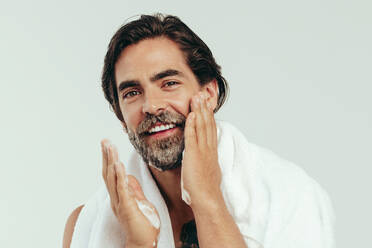 Ein glücklicher weißer Mann ist stolz auf seine Pflegeroutine und trägt sorgfältig Rasierschaum auf seinen Bart auf. Mit ruhiger Hand und konzentriertem Blick bereitet er sein Gesicht auf eine gründliche Rasur in einem Studio vor. - JLPSF29973